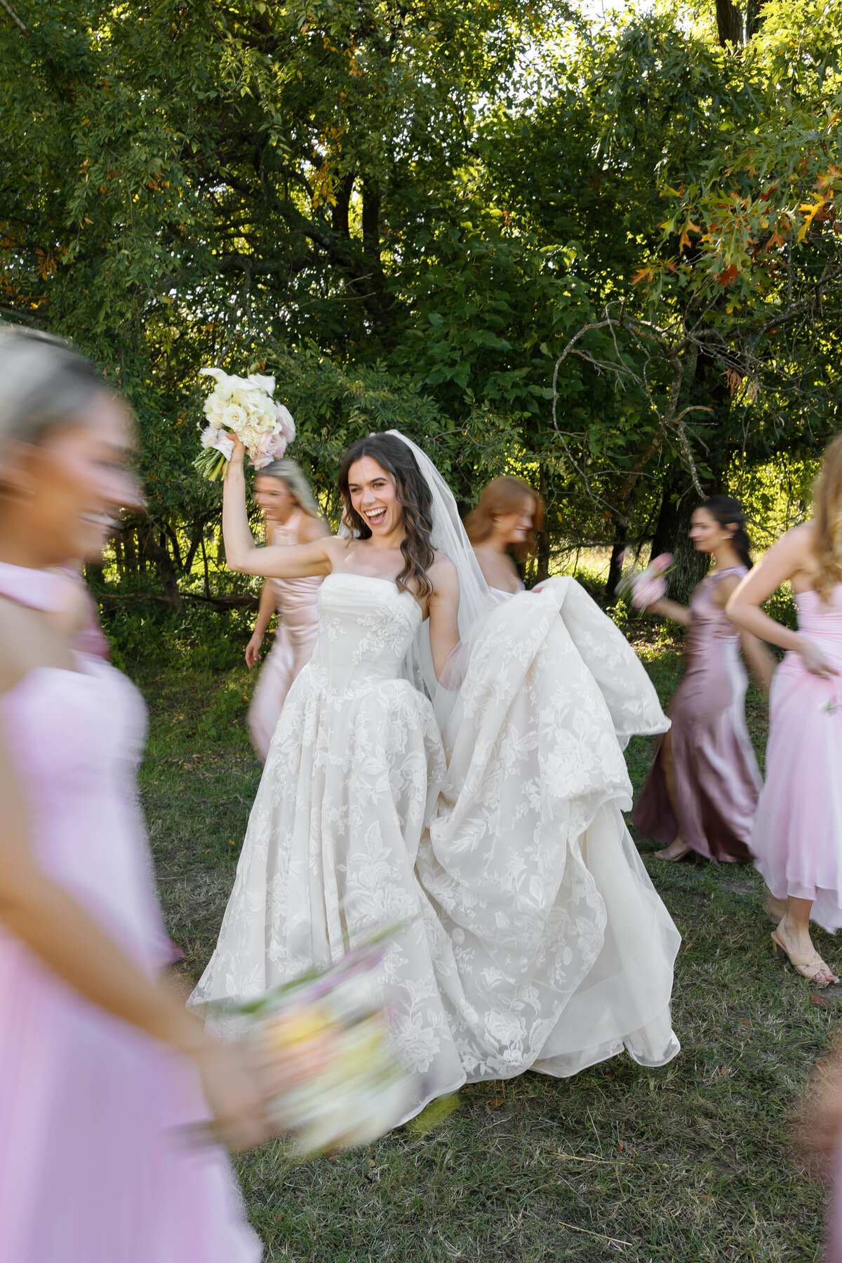 Bride celebrating with bridesmaids as they walk outdoors, holding a bouquet of ivory and blush flowers including roses and calla lilies in a joyful wedding moment.