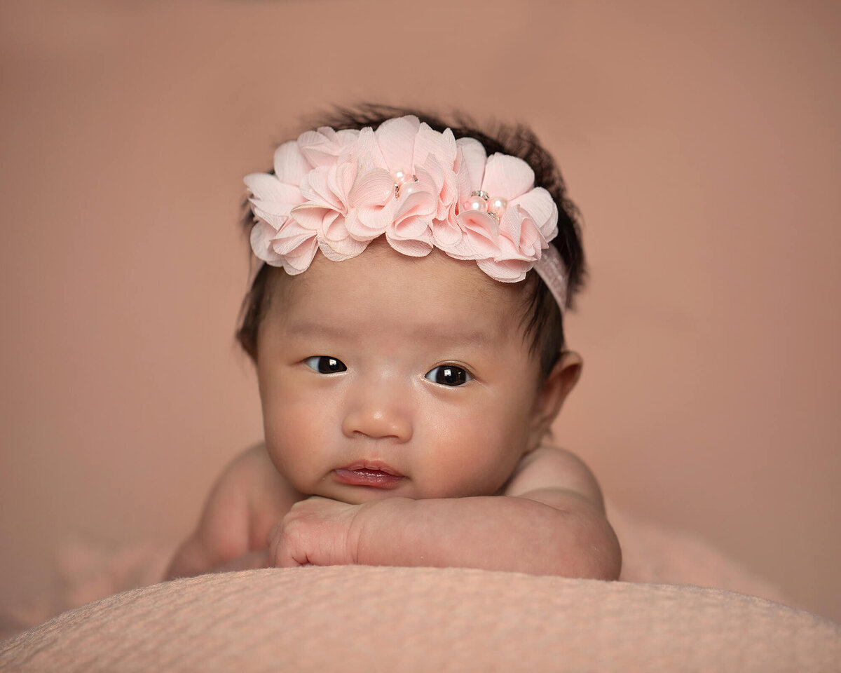 Newborn baby girl reating her chin on her arms as she looks forward wide eyed. Lovely peach headband and background.