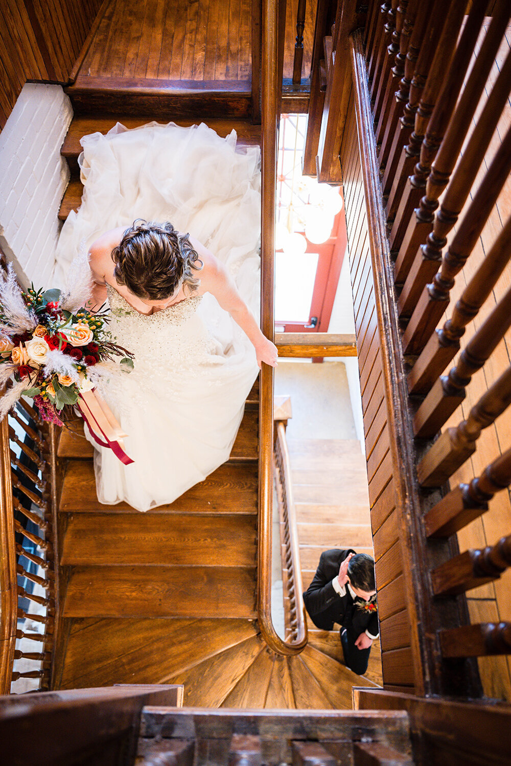 A bride descends the stairs holding onto the railing with one hand and her wedding bouquet in the other while the groom ascends the stairs fixing his hair as the two meet in the center of the staircase for a first look on their elopement day in Virginia.