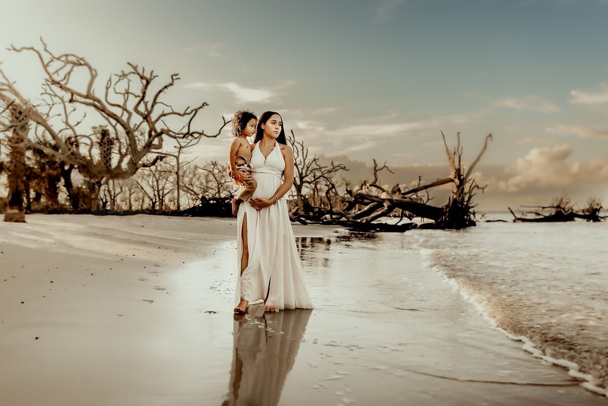 Woman in a white dress holds a child on a beach with driftwood trees, against a backdrop of a cloudy sky.