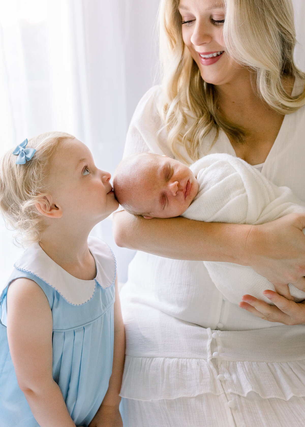 Studio newborn session in white studio. Sibling kissing swaddled newborn in mom's arms. Studio located and captured by  Brandon, MS Newborn  Photographer