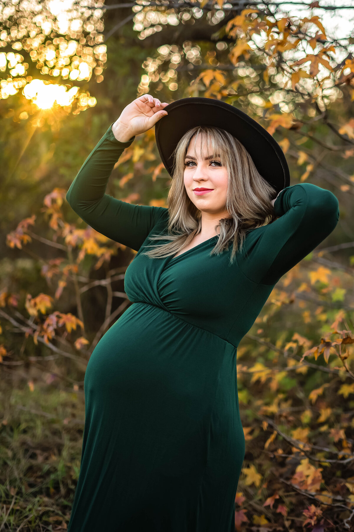 Pregnant woman in a long green dress at sunset during the fall season holding a hat