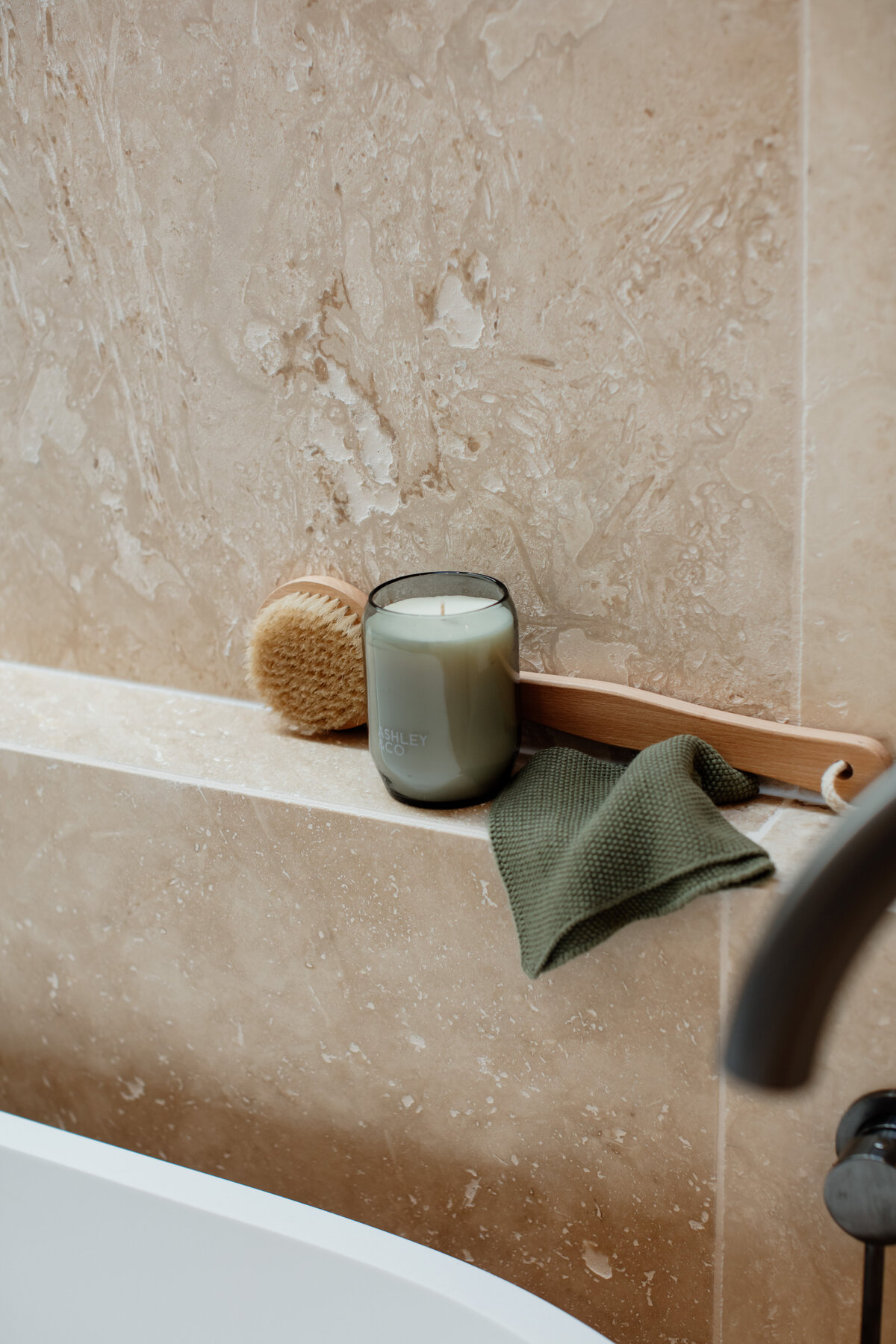 A candle, brush, and cloth are elegantly arranged on a beige marble bathtub ledge, reflecting the bespoke interior design Newcastle NSW is renowned for.