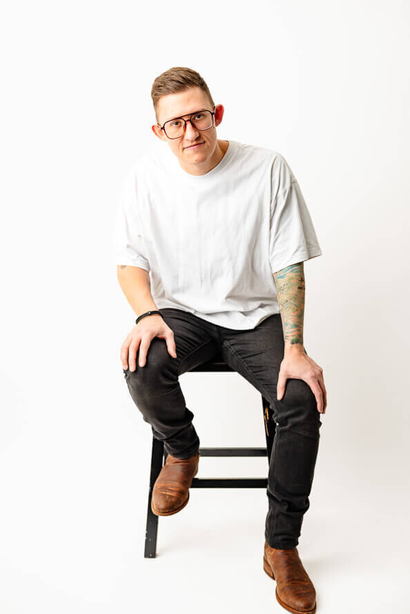 Man in white shirt and black pants sits on stool and leans on legs in studio of Angela Marie Media.