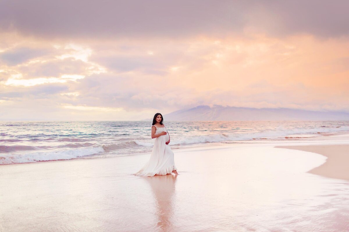 Woman with long black hair holds belly while wearing white maternity dress for her Maui sunset babymoon session
