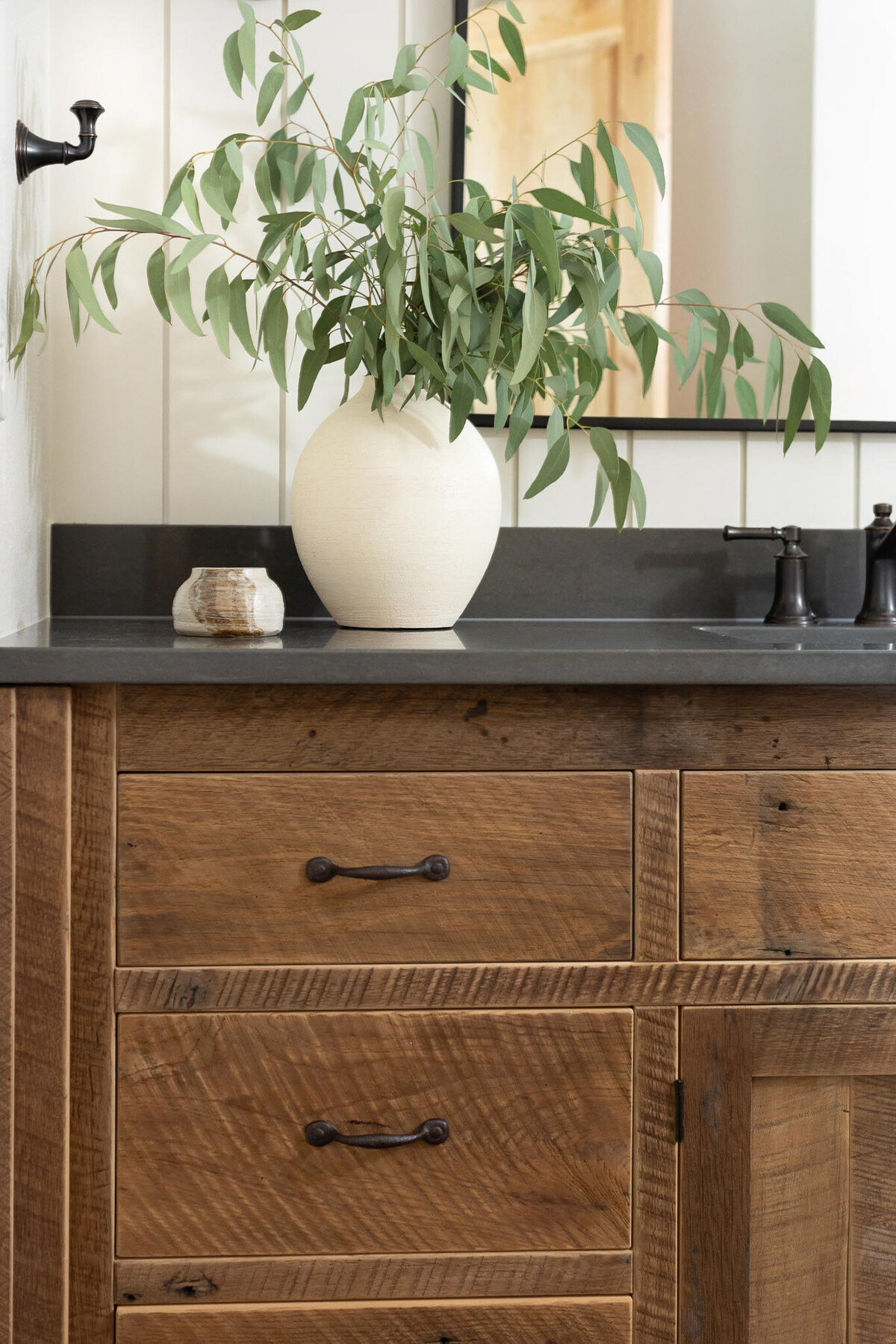 Large rustic wood vanity with lots of drawers, and dark grey countertops. Wall behind the vanity is off-white shiplap with a large arched mirror and two oil rubbed bronze sconces on either side. A widespread oil rubbed bronze faucet at the sink with a large beige vase with green florals to the left of the sink. To the right of the sink a jar of hand soap and a small beige dish. Large rustic black square floor tiles with brown grout.