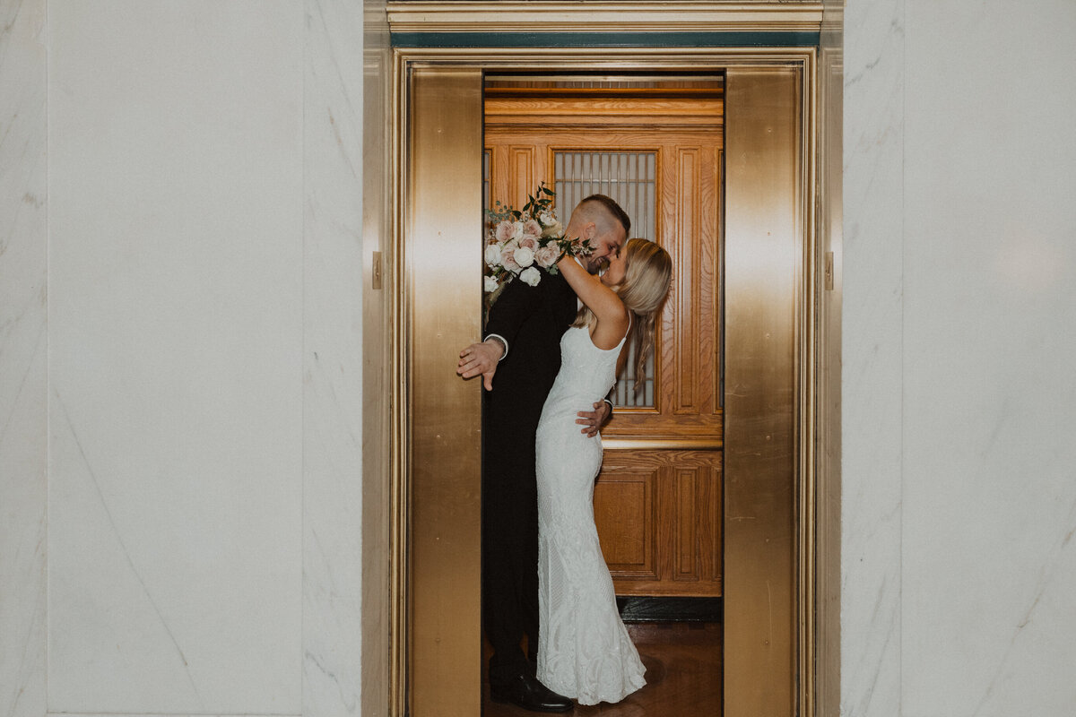 bride and groom kissing in elevator while groom holds the door open