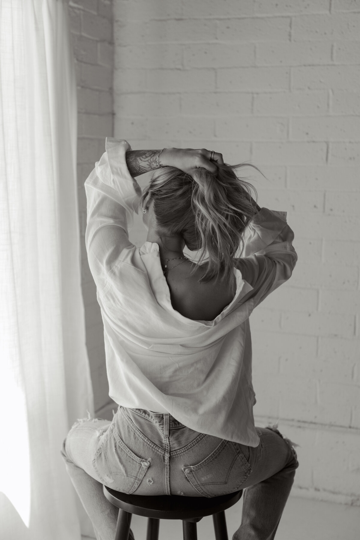 Black and white branding portrait of a woman on a stool pulling her hair up with her back to the camera, wearing a white shirt and jeans — editorial image by Dani Lacey in Greenville SC.