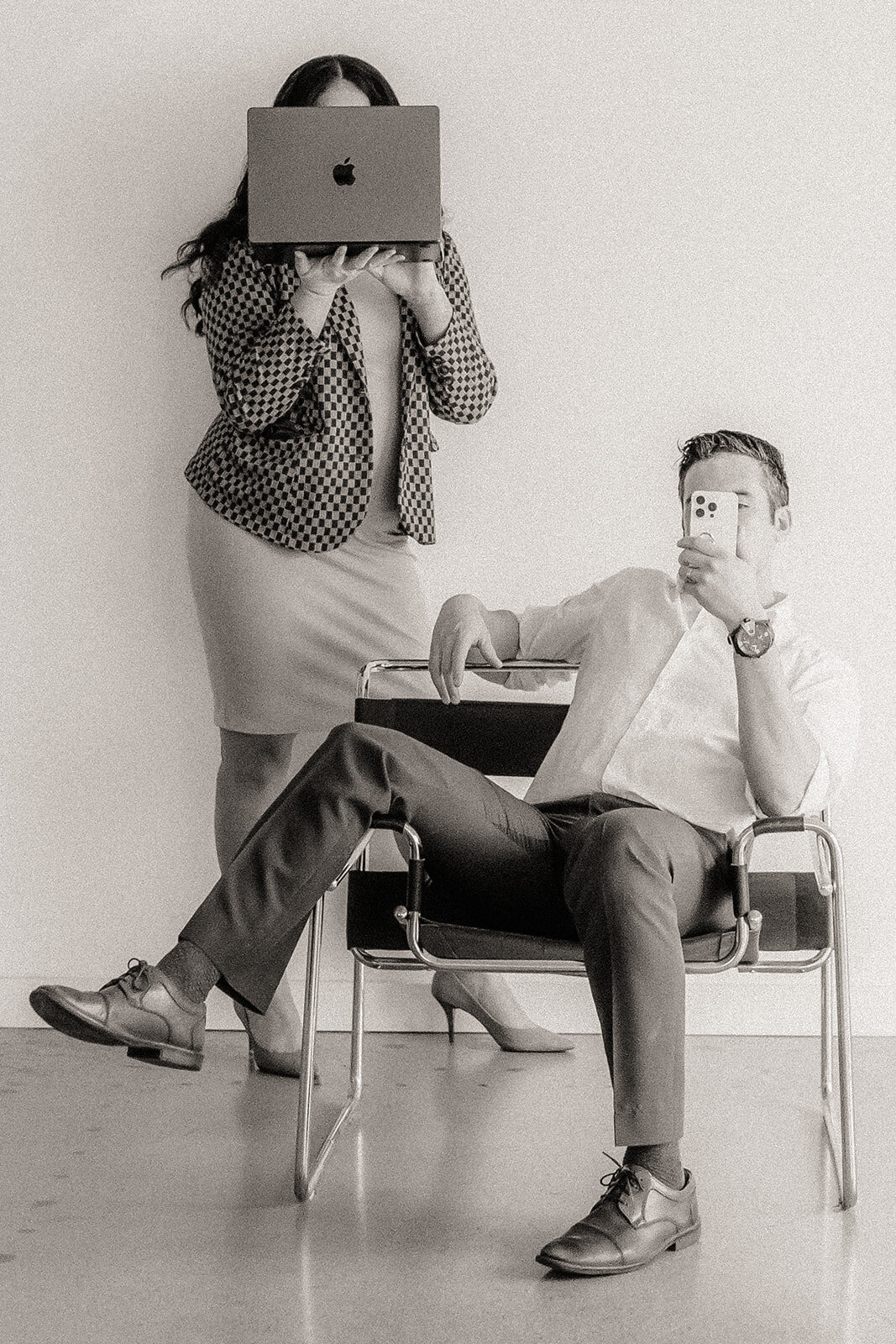A man holds a phone in a chair while a woman stands behind on a laptop