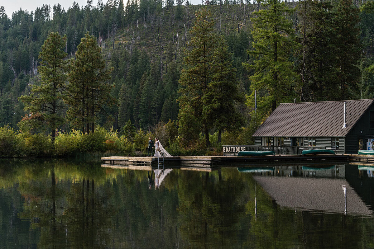 A bride and groom stand on a dock with the lake in the foreground and tress and a cabin in the background.