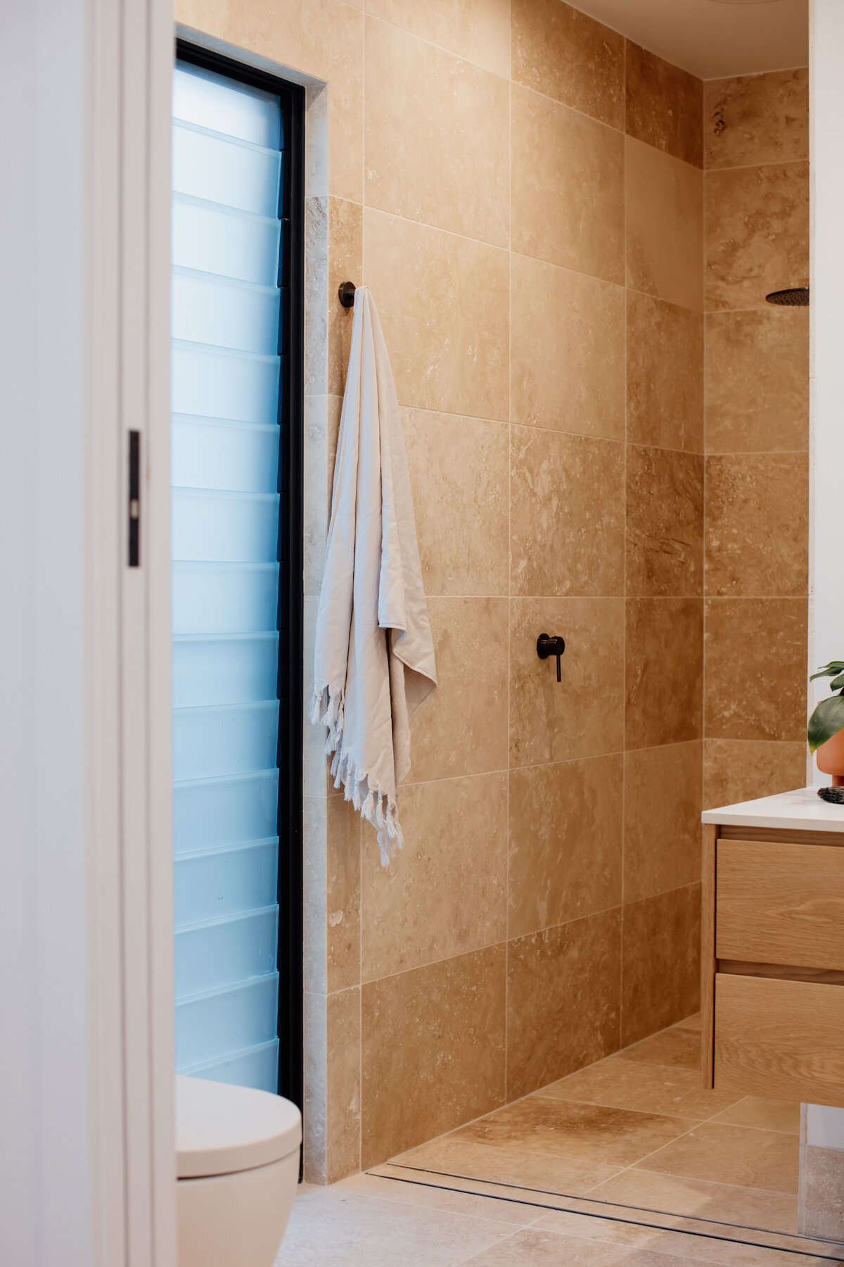 Modern bathroom featuring a beige tiled shower, a hanging towel, frosted window, and wooden vanity, crafted through bespoke interior design in Newcastle NSW.