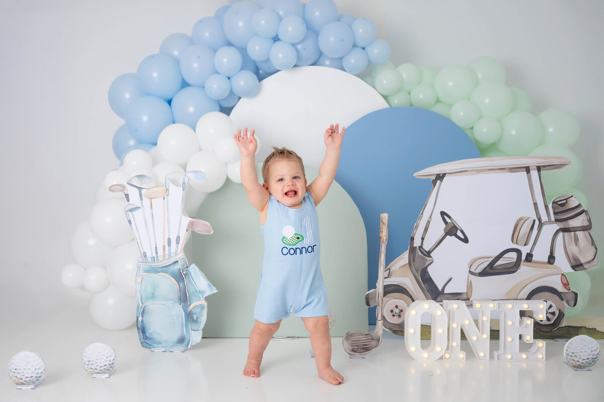 Baby boy in a blue golf-themed romper throws his hands in the air excitedly.