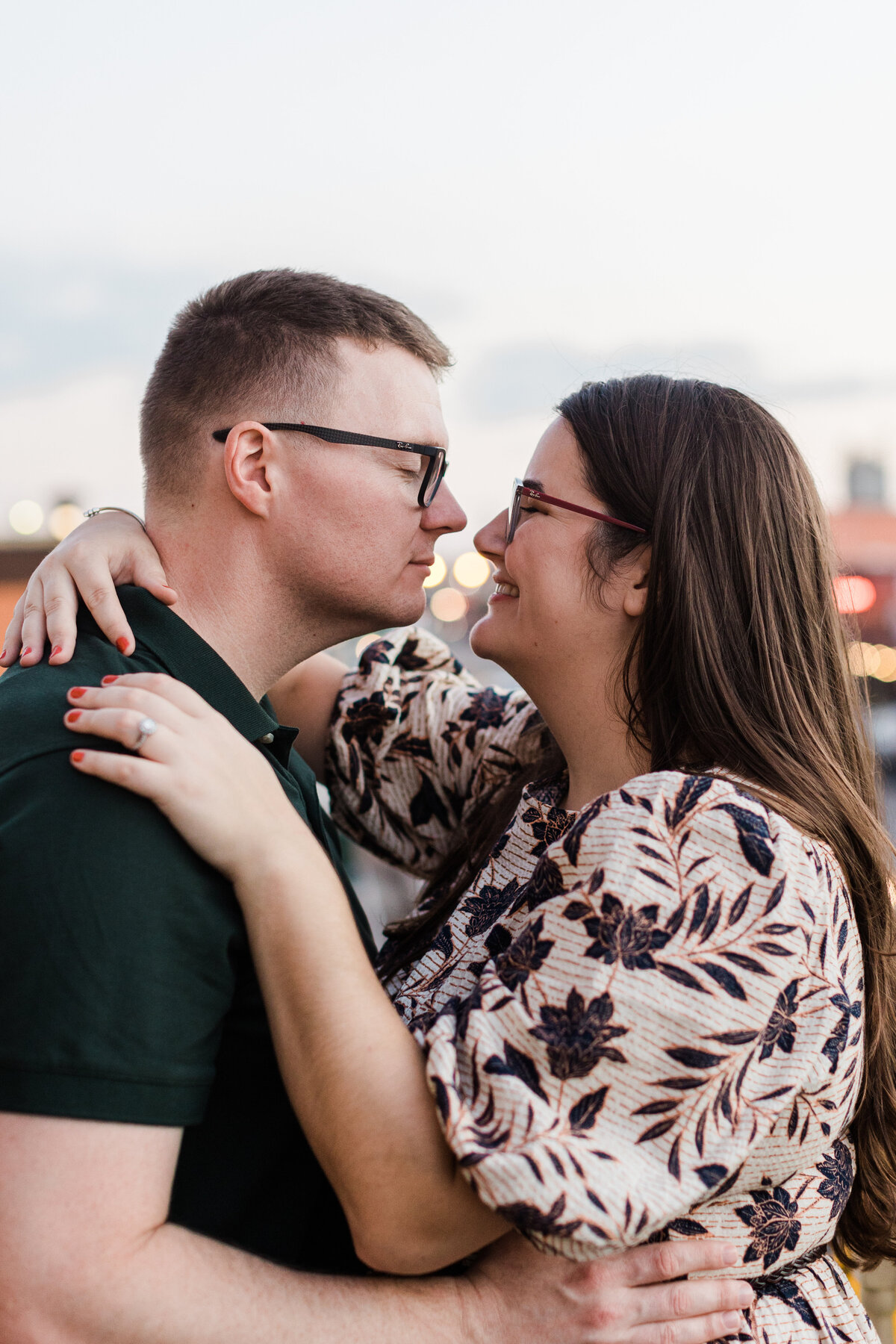 A couple holding each other close and preparing to kiss during their engagement session in Grapevine, Texas. The woman on the right is wearing a white dress covered in dark flowers and glasses. Her left arm is placed on the man's shoulder, so her engagement ring can be seen. The man on the left is wearing a dark green shirt and glasses.
