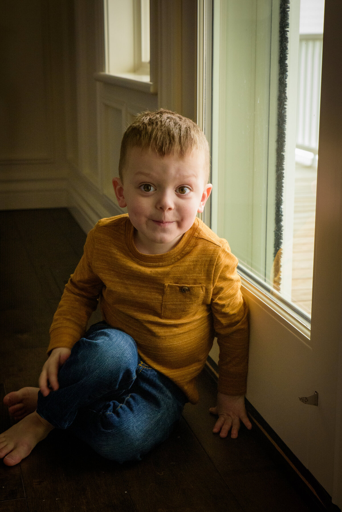 Natural light portrait of young boy wearing mustard yellow shirt sitting next to window in his home in Green Bay, Wisconsin.