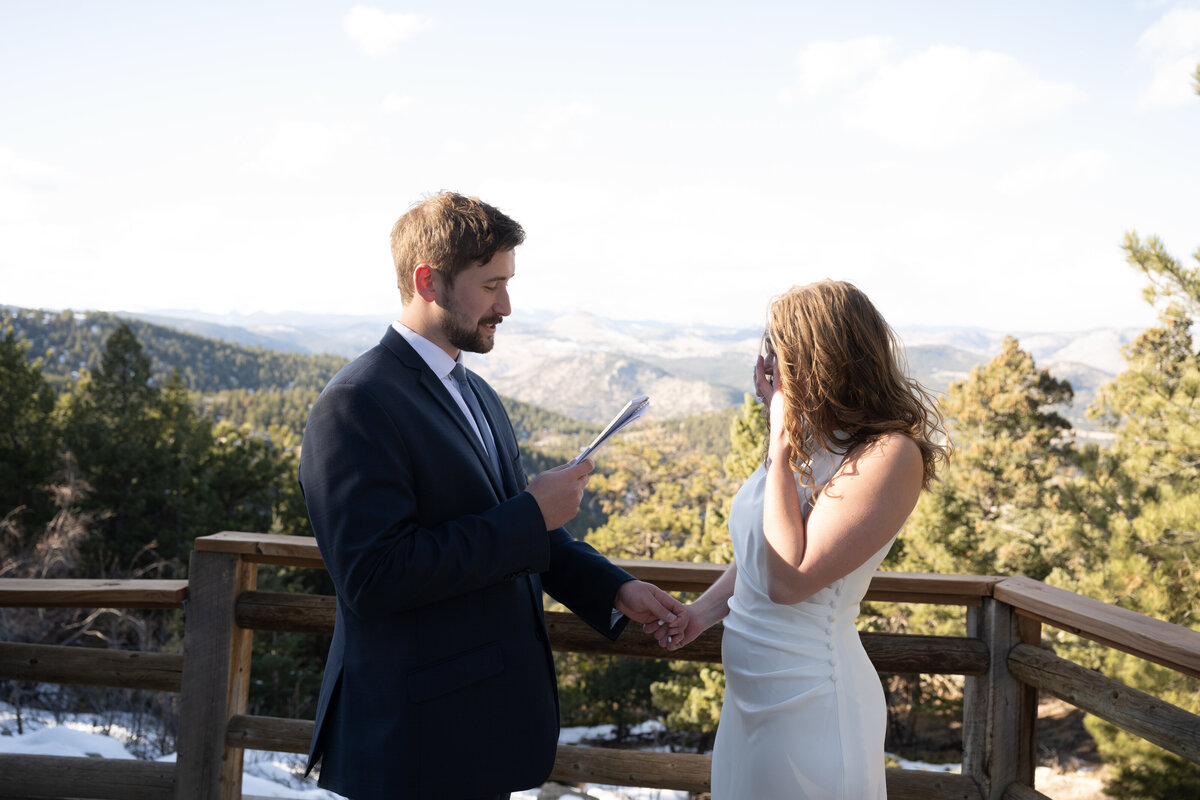 A heartfelt moment unfolds as a couple exchanges vows against a breathtaking mountain backdrop. The groom, dressed in a navy-blue suit, holds a piece of paper, reading his promises with sincerity. The bride, in a simple yet elegant white gown, listens intently, her face illuminated by the sunlight filtering through the trees. Their hands are clasped together, symbolizing the love and commitment they share. The rustic wooden railing and snow-dusted ground add to the natural beauty of their intimate ceremony, making this a timeless and emotional capture of their special day.