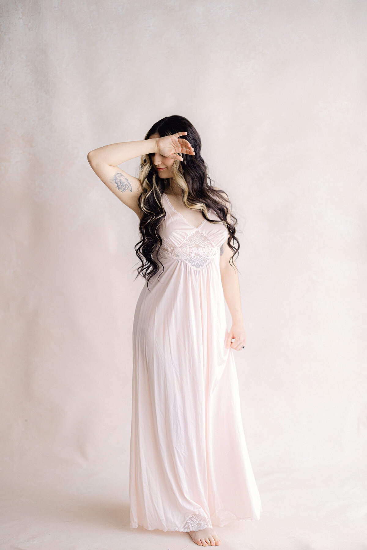 Woman with long brown hair in a silk pink nightgown places her hand over her eyes during a session with Indianapolis boudoir photographer, Brittney Lear.