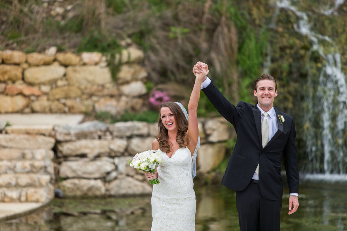 Bride and groom holding hands and lifting them up into the air after saying "I do" at their Hill Country wedding venue Hidden Falls