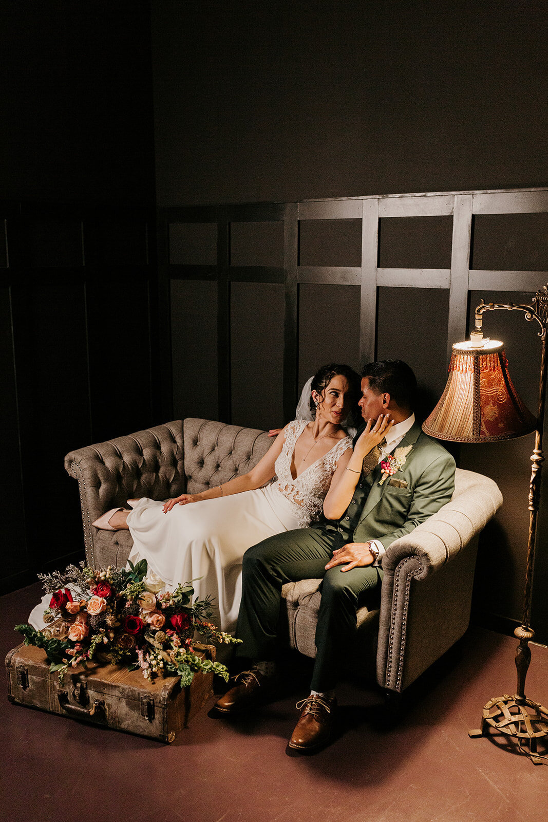 The bride and groom share an intimate moment on a vintage couch under warm light, with a floral bouquet and a vintage suitcase in front of them.