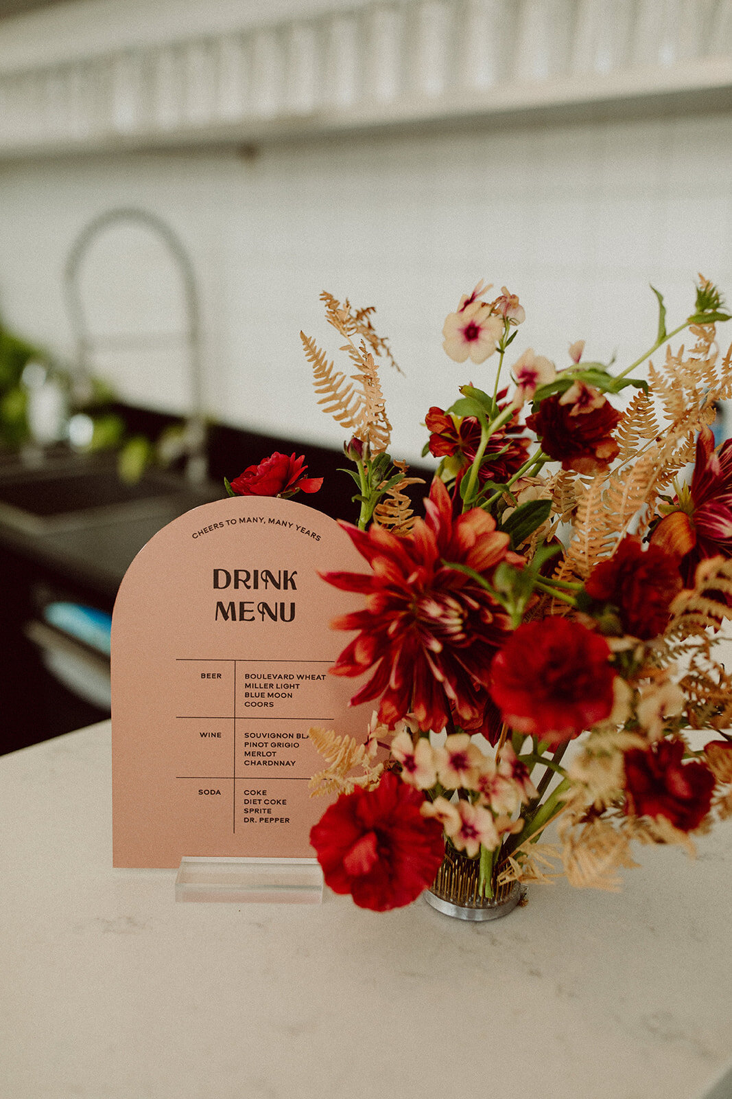 Rounded peach drink menu with black font on clear place card holder atop a white table with wine-colored flowers.