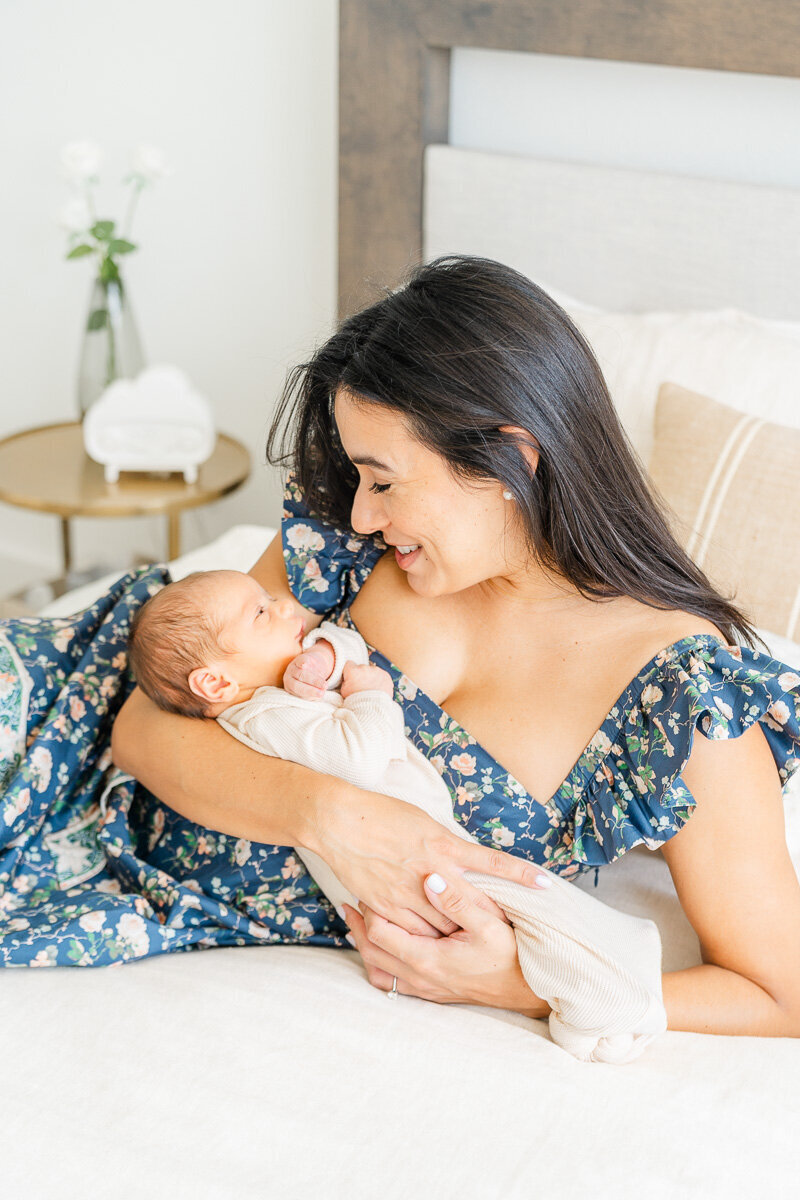 a mother sits on a bed in her Leander home while holding her newborn baby and smiling at him.