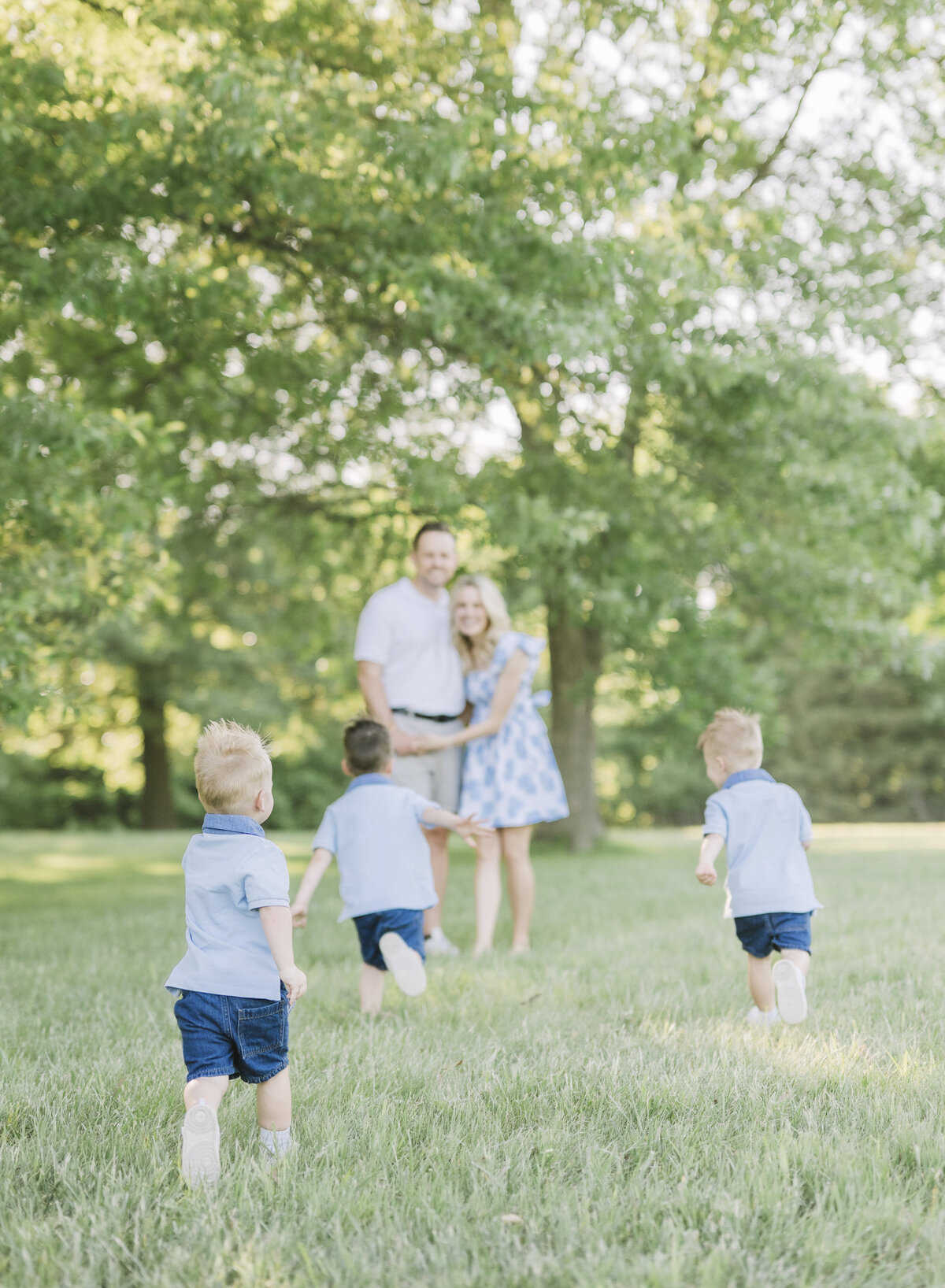 Parents hug while three toddlers run through a sunny grassy field in a matching blue outfit set.