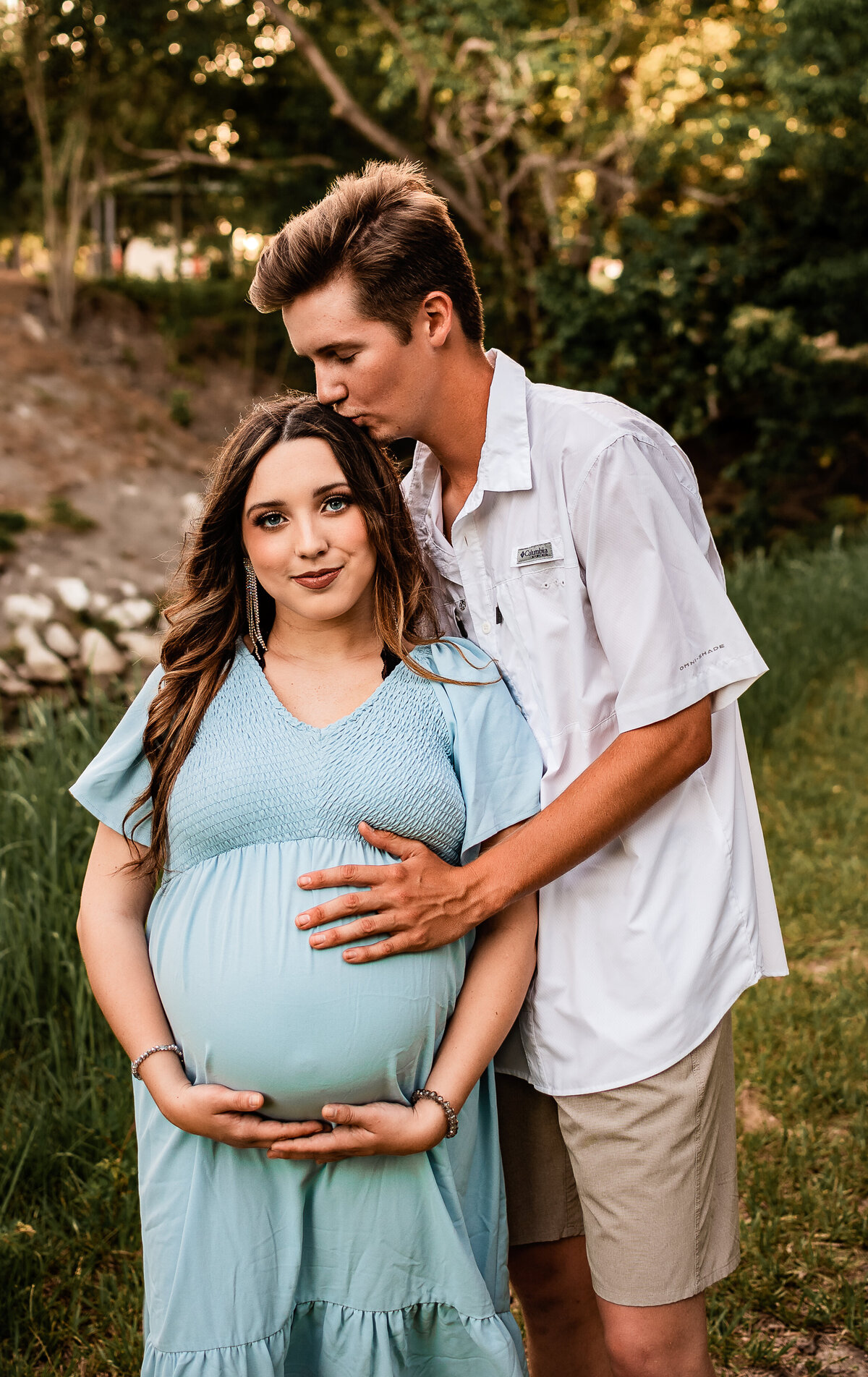 An expecting couple standing in front of a creek hold mom to be's belly.
