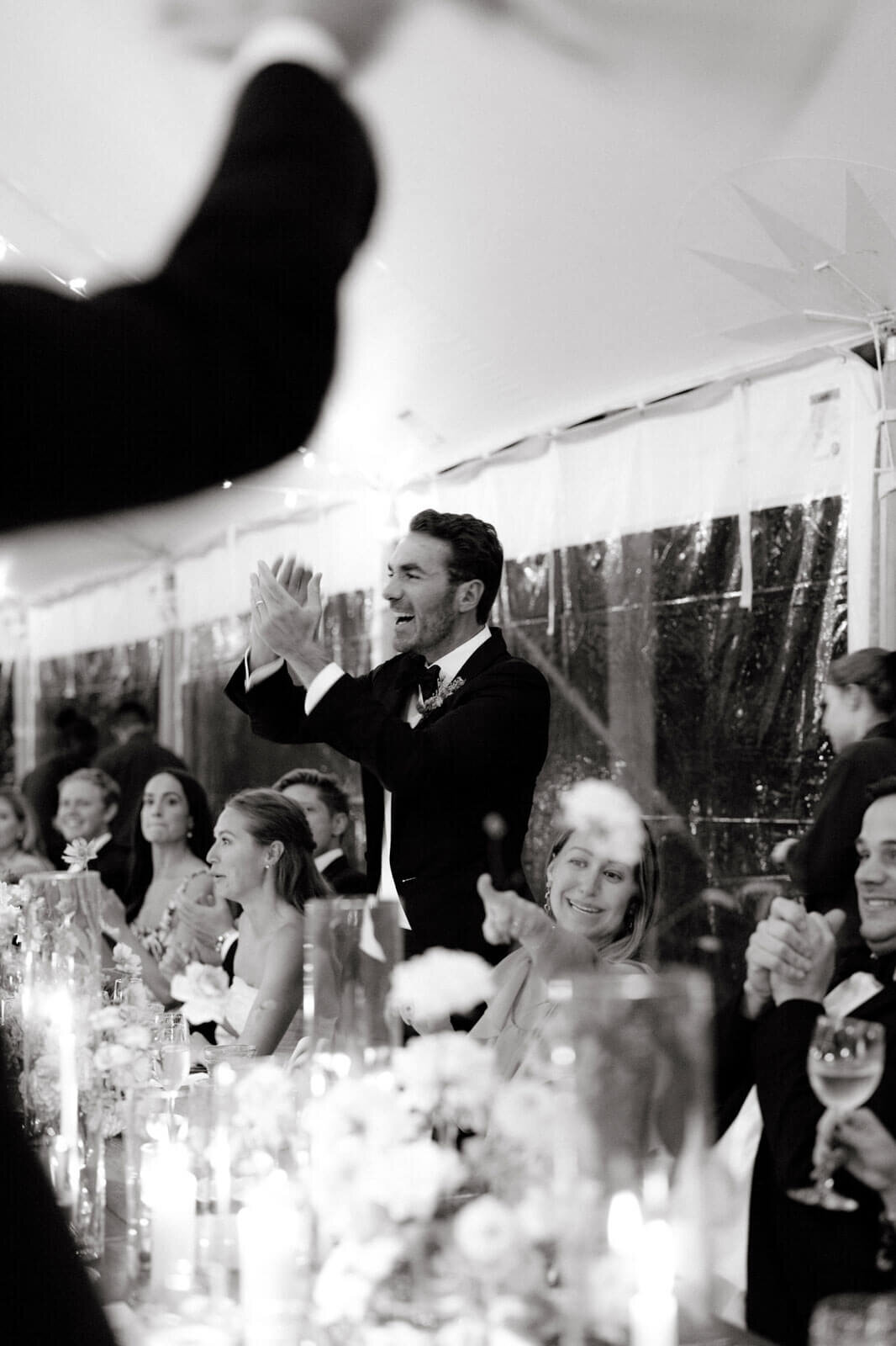 The groom is happily standing, laughing, and clapping at a wedding reception at The Ausable Club, NY. Image by Jenny Fu Studio