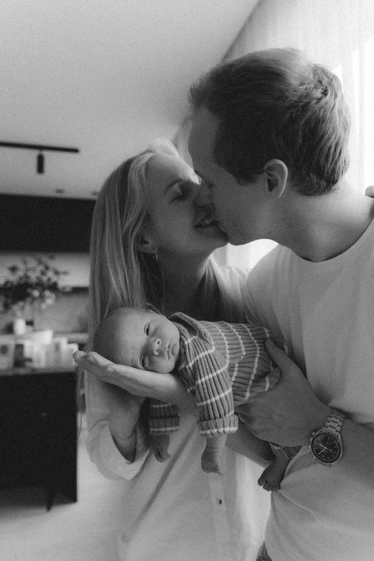 Zwart-wit foto van newborn baby op de arm van de papa en mama geeft een kus.