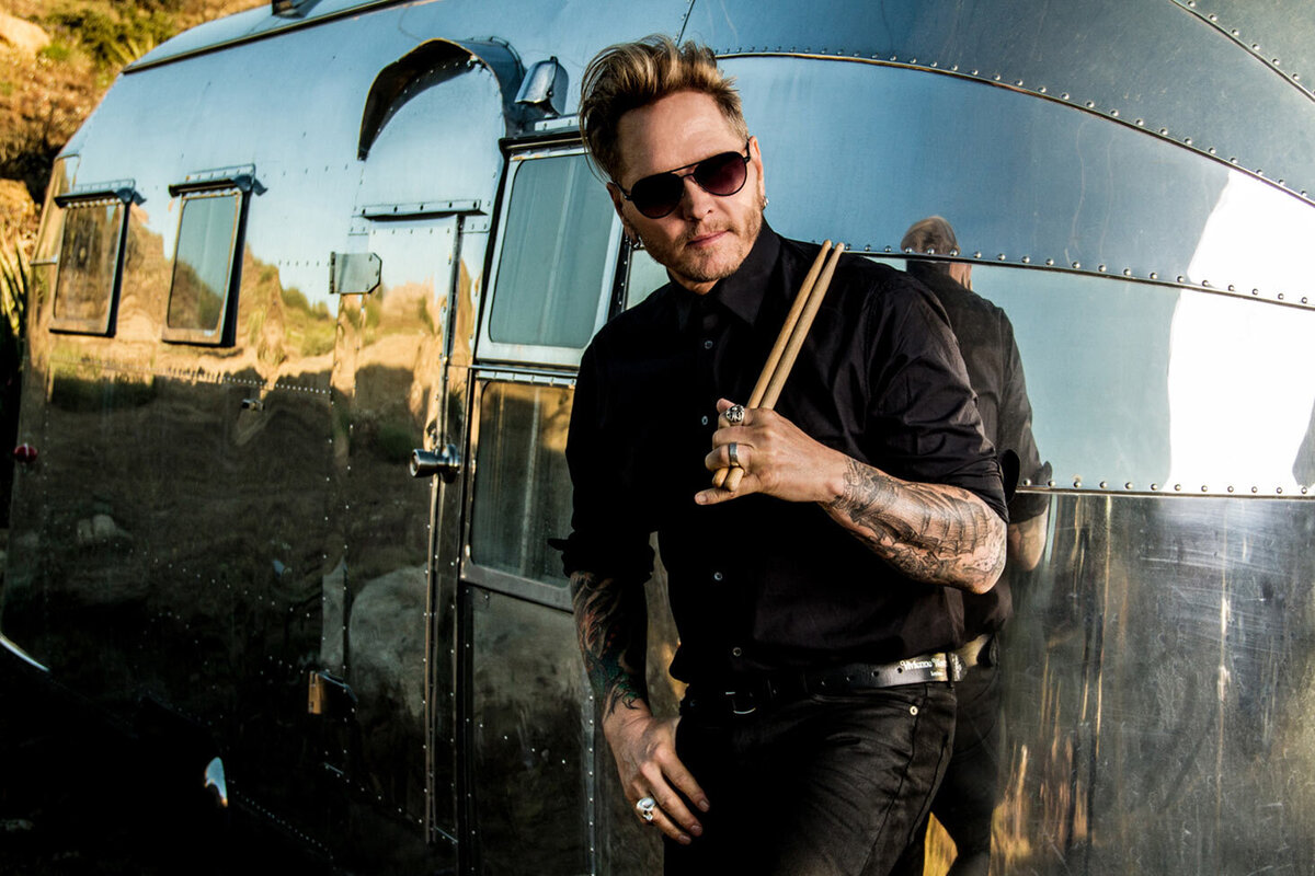 Matt Sorum musician portrait standing against silver airstream trailer holding drumsticks that are leaned on his shoulder other hand hanging off his pants pocket