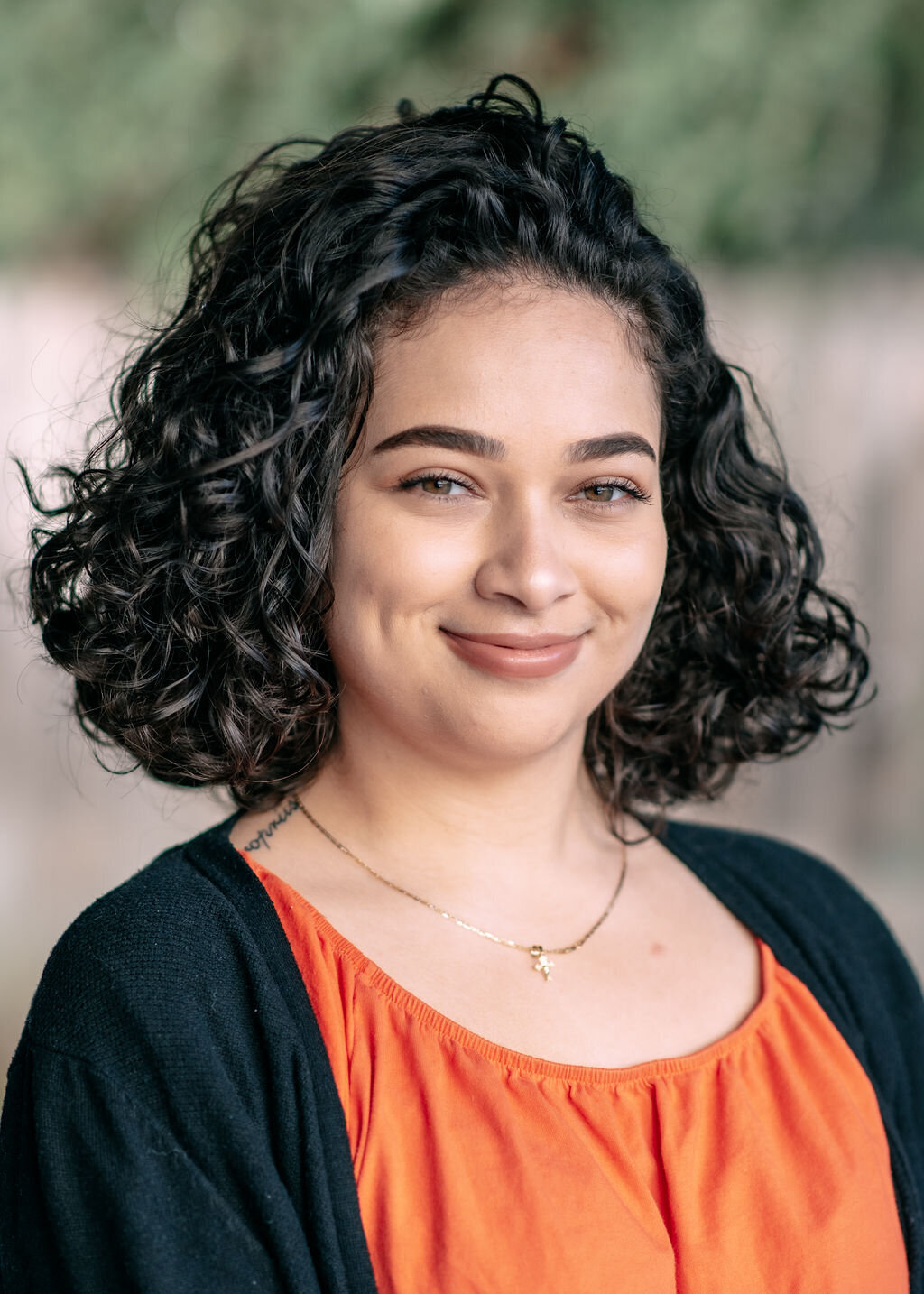 Woman with black curly hair wearing an orange top and a black cardigan stands outside for a headshot with Lisa Winner Photography