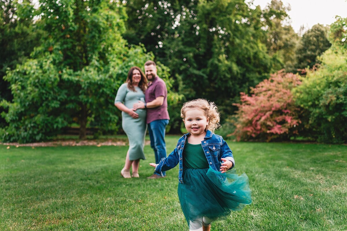 daughter running with pregnant mom and dad in background at park