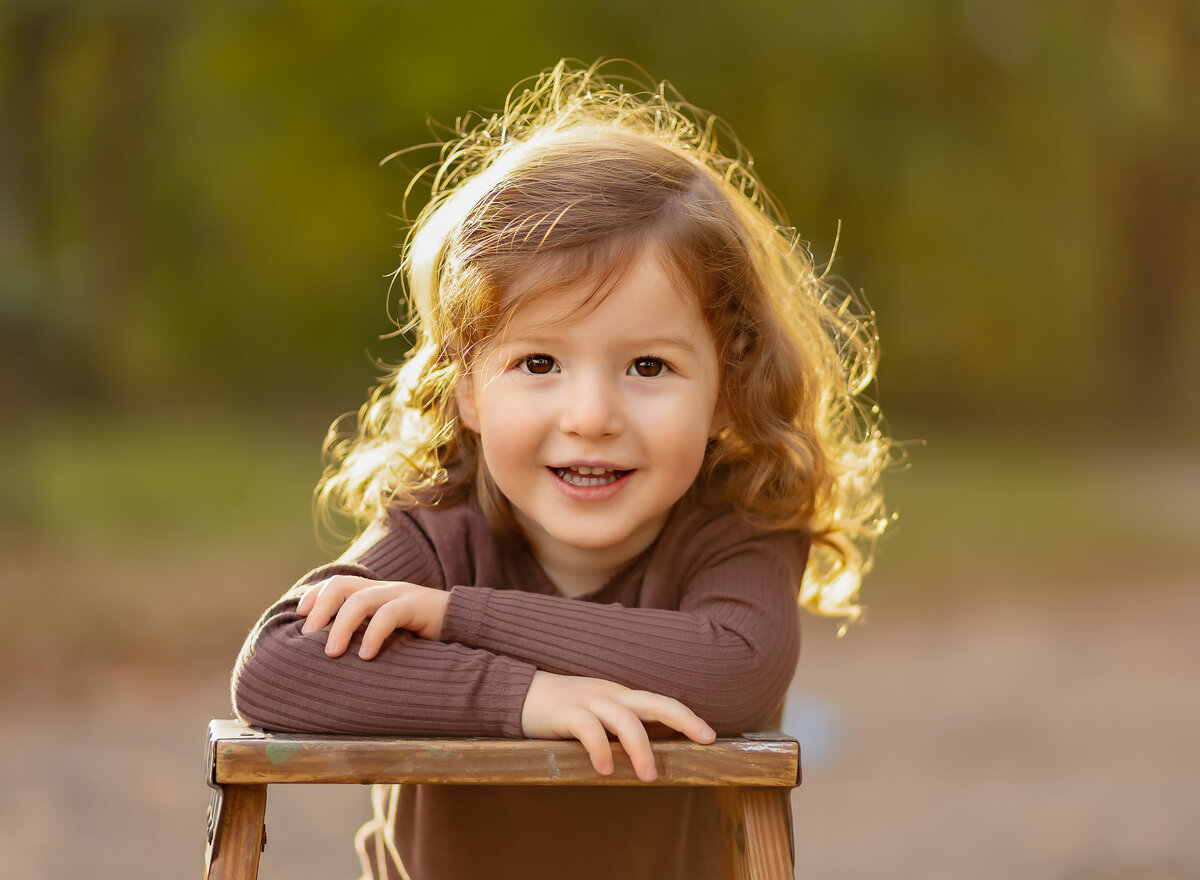 Little boy sits on a stool at golden hour for his brooklyn ny family photoshoot. His hands are folded on the stool and he is smiling at the camera. Captured by brooklyn family photographer Chaya Bornstein.