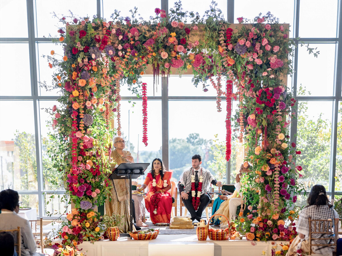 A vibrant Indian wedding scene with a floral arch framing a couple seated on stage. The setup includes colorful flowers and greenery. A speaker stands to the left, and attendees are visible in the foreground. Sunlight streams through large windows behind them.