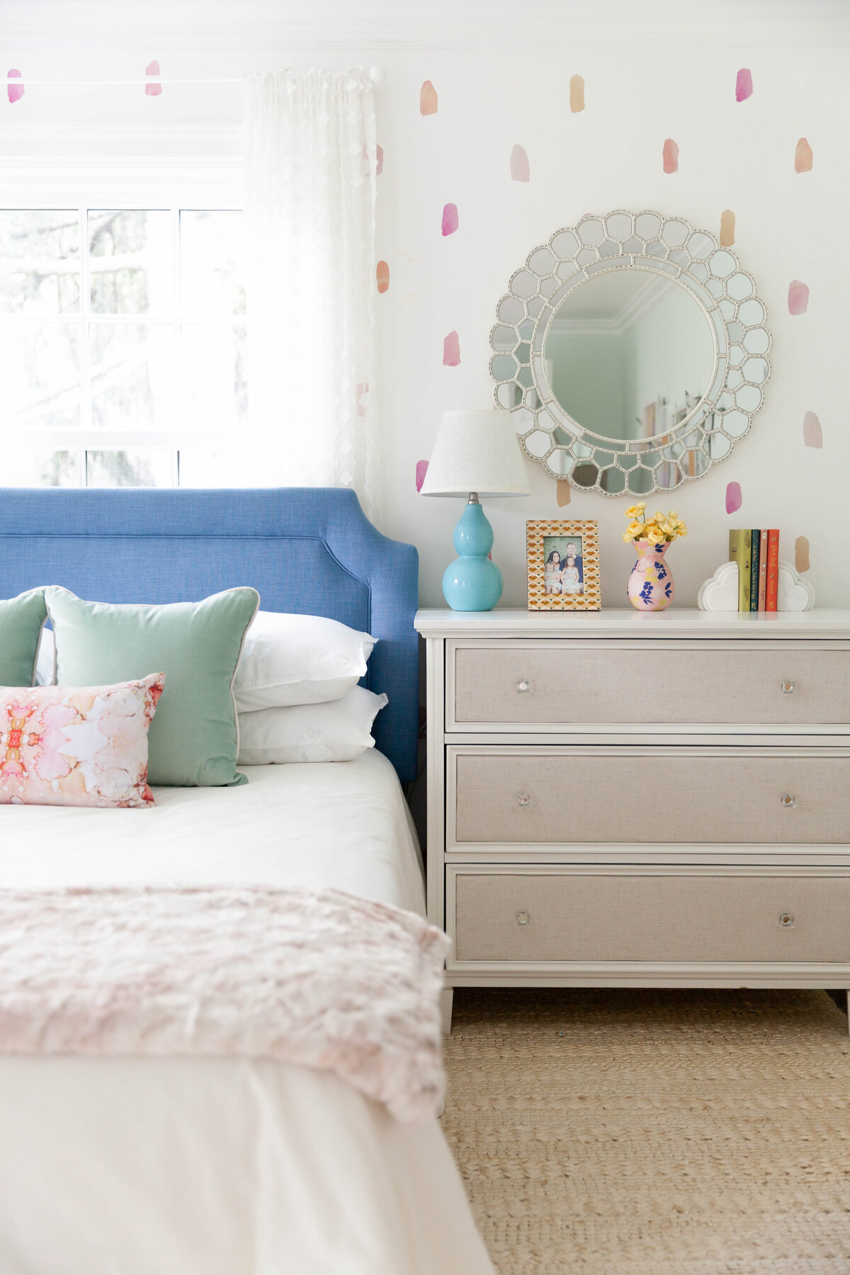 Detail of a luxury kids’ bedroom designed by Deni + Dove Interiors in Montclair, NJ. Featuring a custom blue upholstered headboard, soft layered bedding, whimsical pastel wall detail, and elegant dresser styling, this space offers a perfect blend of charm and sophistication for modern family living in Essex County.