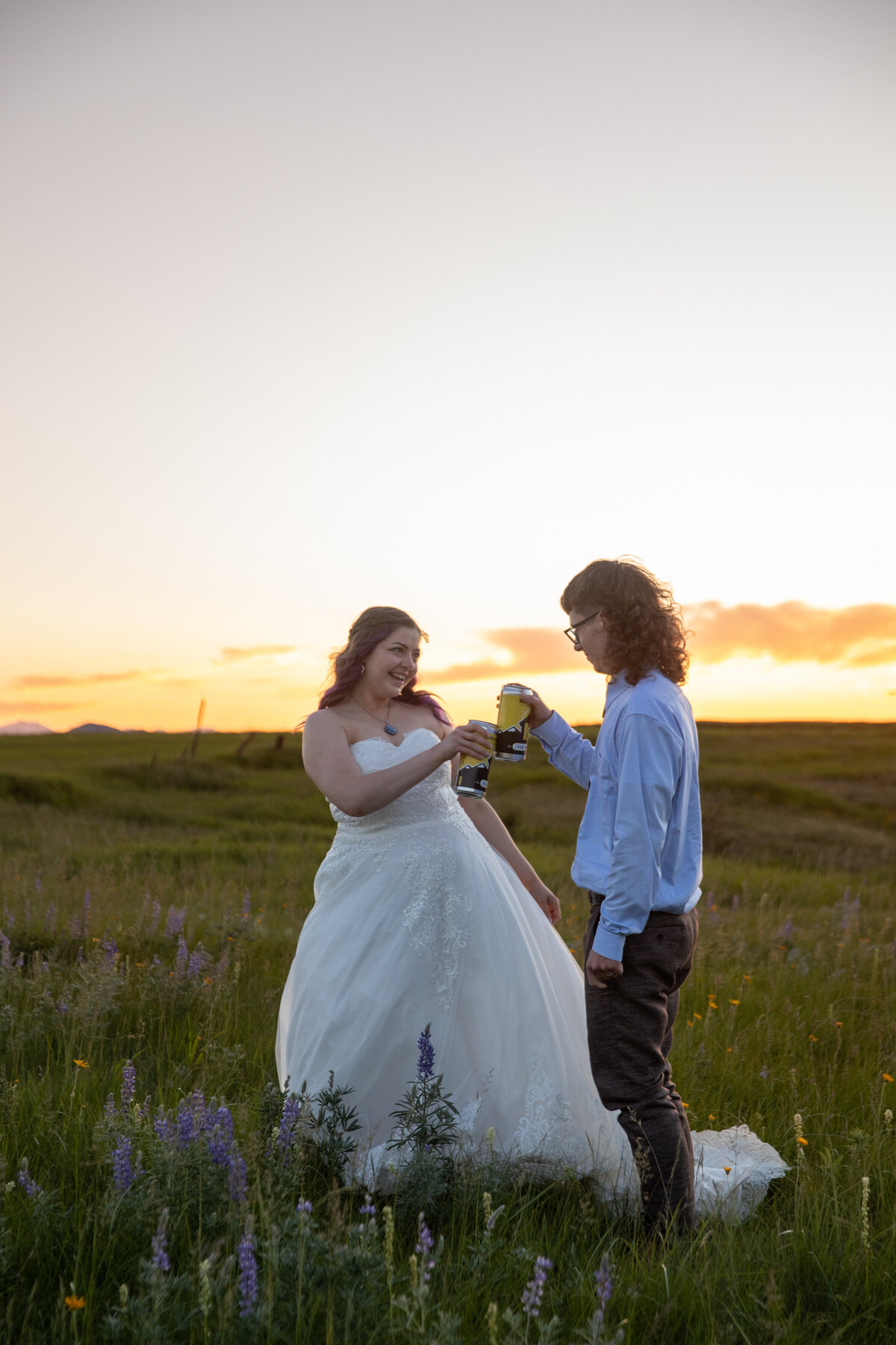 A bride and groom stand in a meadow of wildflowers, cheersing their beer cans on their elopement day in Montana.