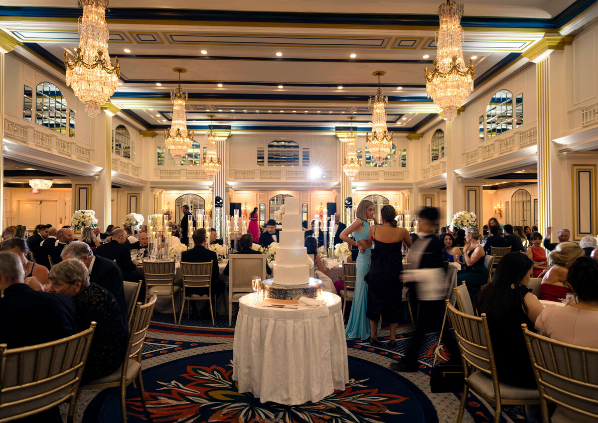 A lavish ballroom filled with elegantly dressed guests seated at tables. A multi-tiered wedding cake is displayed on a round table in the center. Chandeliers hang from the ceiling, and large mirrors adorn the walls, reflecting the opulent setting.