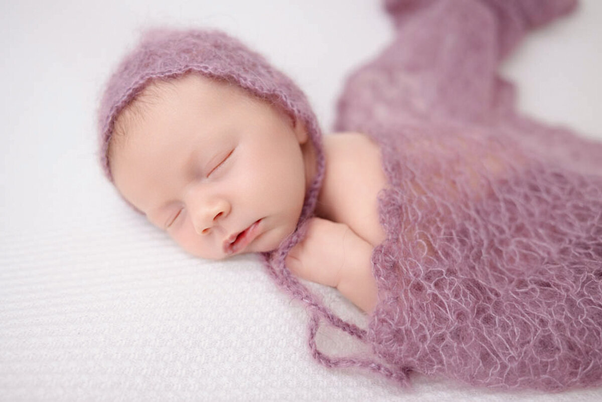 two-week baby girl posed lying on white blanket and wearing a purple bonnet and covered in a lacy purple baby wrap.