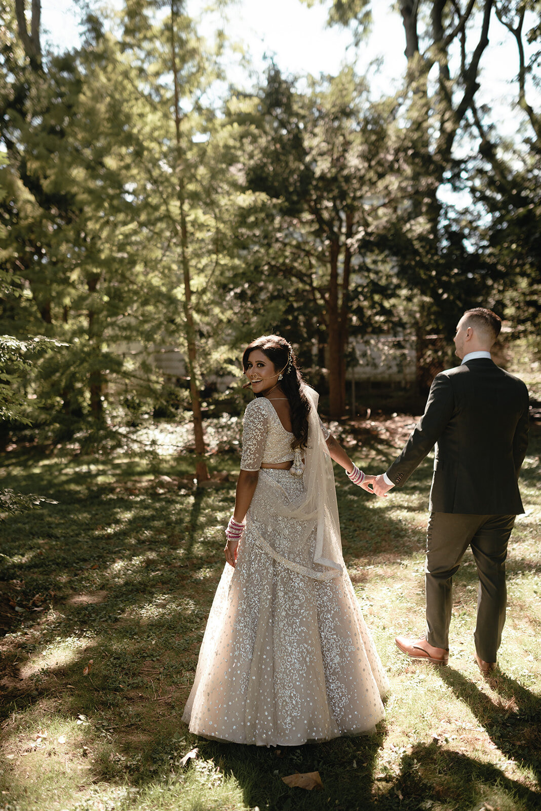 A bride in an embellished champagne-colored lehenga glancing back while holding hands with the groom as they walk through a sunlit forested area.