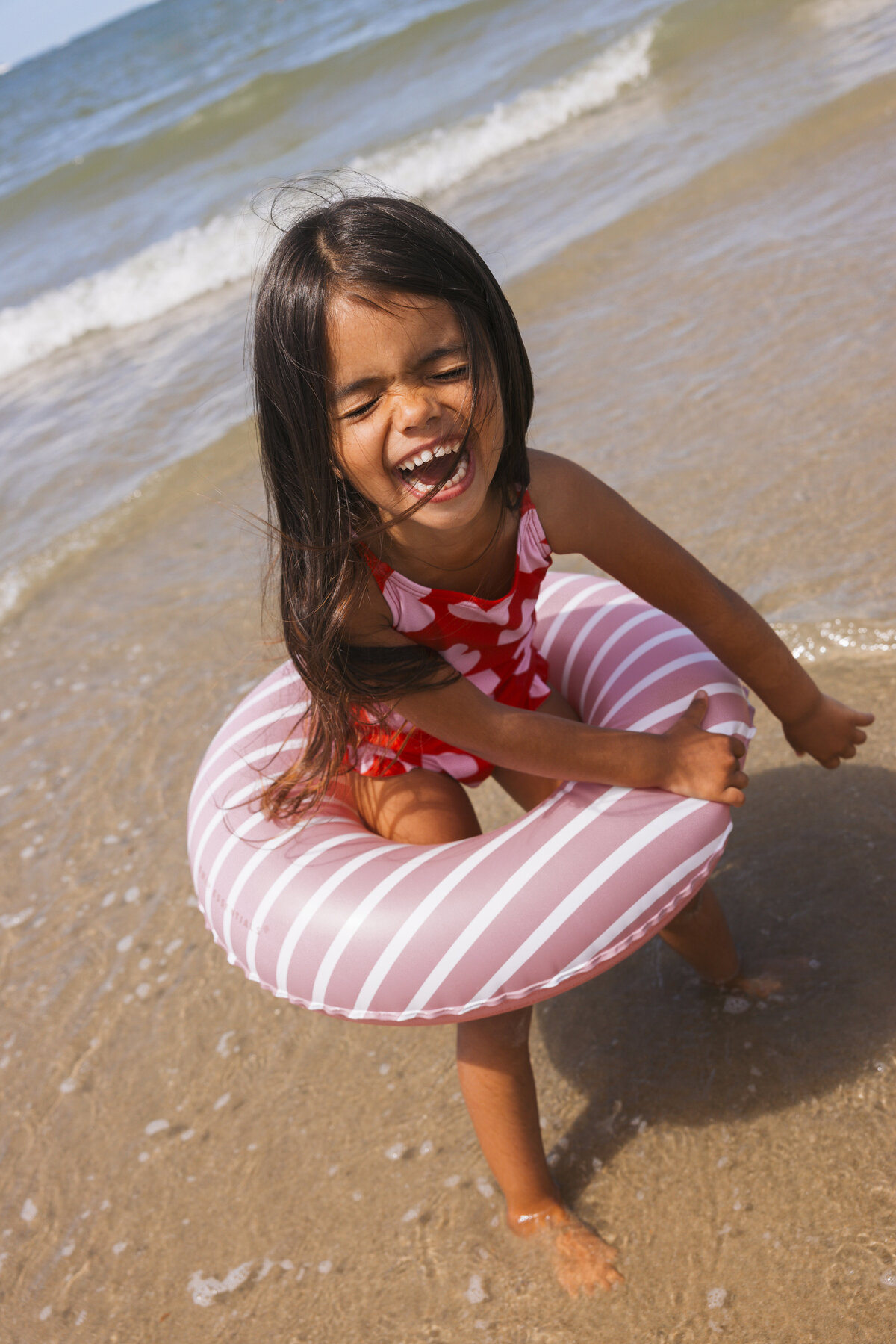 Laughing girl in a red heart-print swimsuit plays in the sea with a pink striped Swim Essentials inflatable ring.
