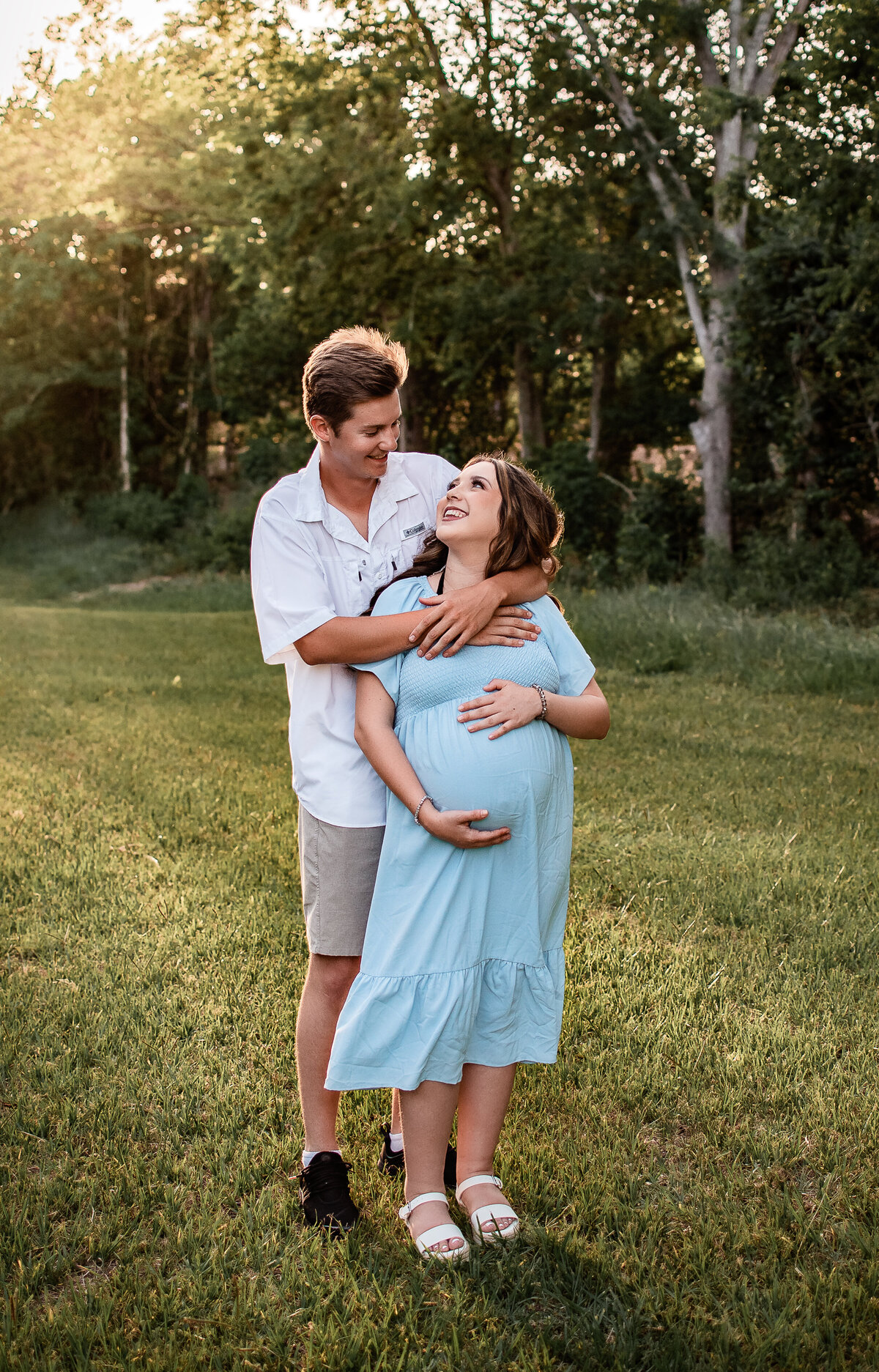 A pregannt couple stands in front of a line of trees and smiles at each other.