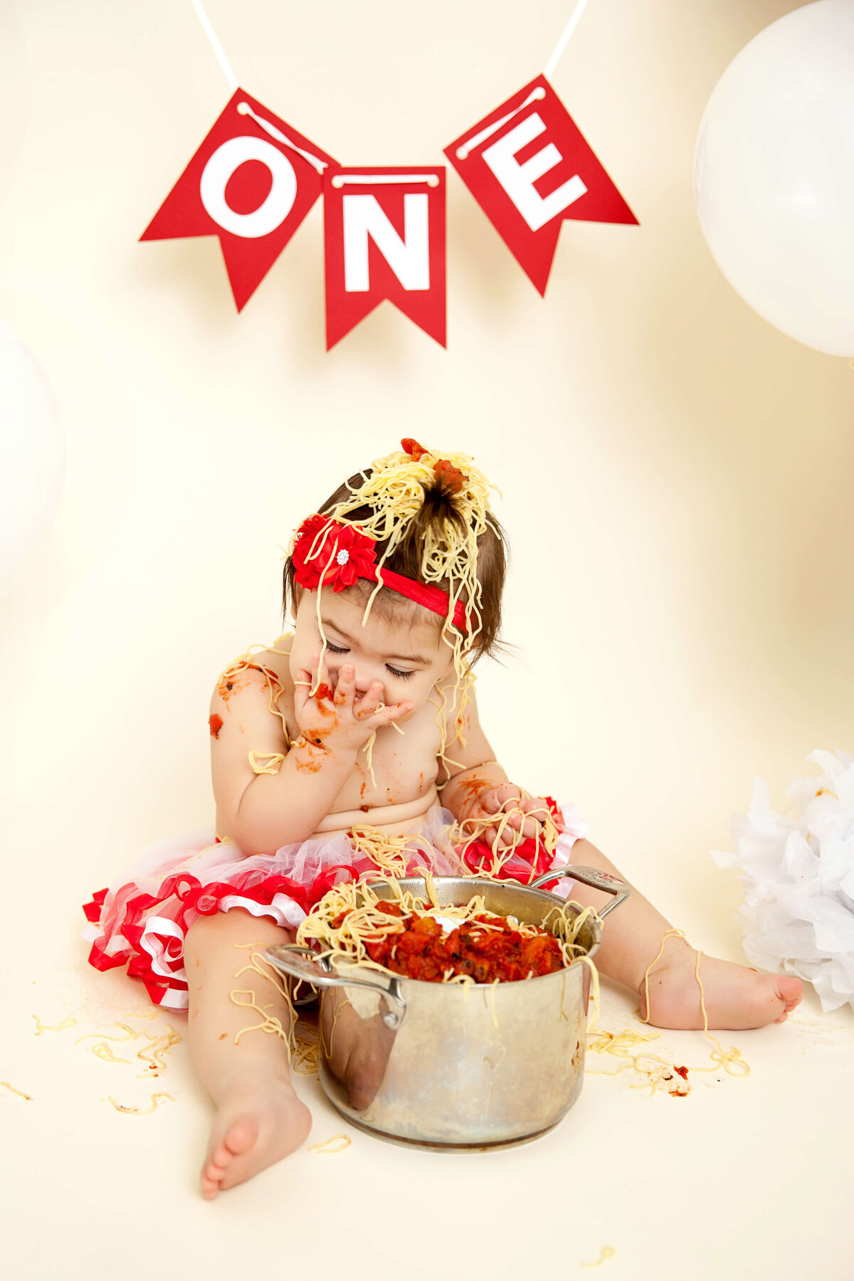 A baby in a red and white outfit sits on the floor, joyfully messy with spaghetti and sauce. A pot of spaghetti is at their feet. Above, a red banner reads "ONE," while white balloons float around—perfect for capturing moments like these with Jacksonville Cake Smash Photography.
