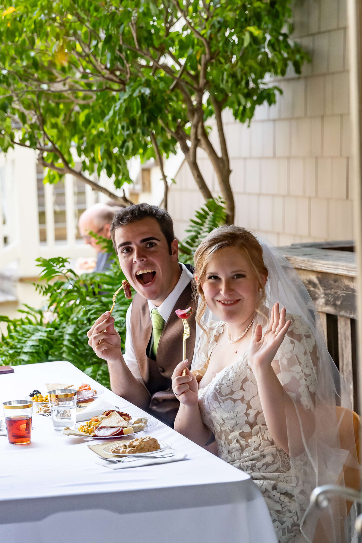 A bride and groom sit at a table outdoors, smiling at the camera while holding forks. The bride waves, wearing a white wedding dress and veil, and the groom wears a suit with a green tie. Plates of food and drinks are on the table.