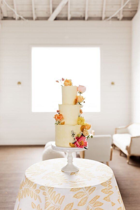 In a modern classy barn-like room, a three tier yellow wedding cake sits on a cocktail table with a patterned yellow and white tablecloth. It is framed by a window with light streaming through, and decorated with fresh flowers in pinks, oranges, and yellows, with candied fruit.