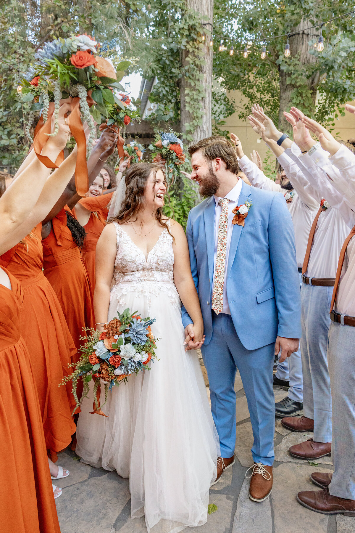 The bride and groom smile at each other while walking through a tunnel of cheering bridesmaids and groomsmen, with vibrant floral bouquets raised in celebration. The bride wears a lace gown and the groom is in a light blue suit.