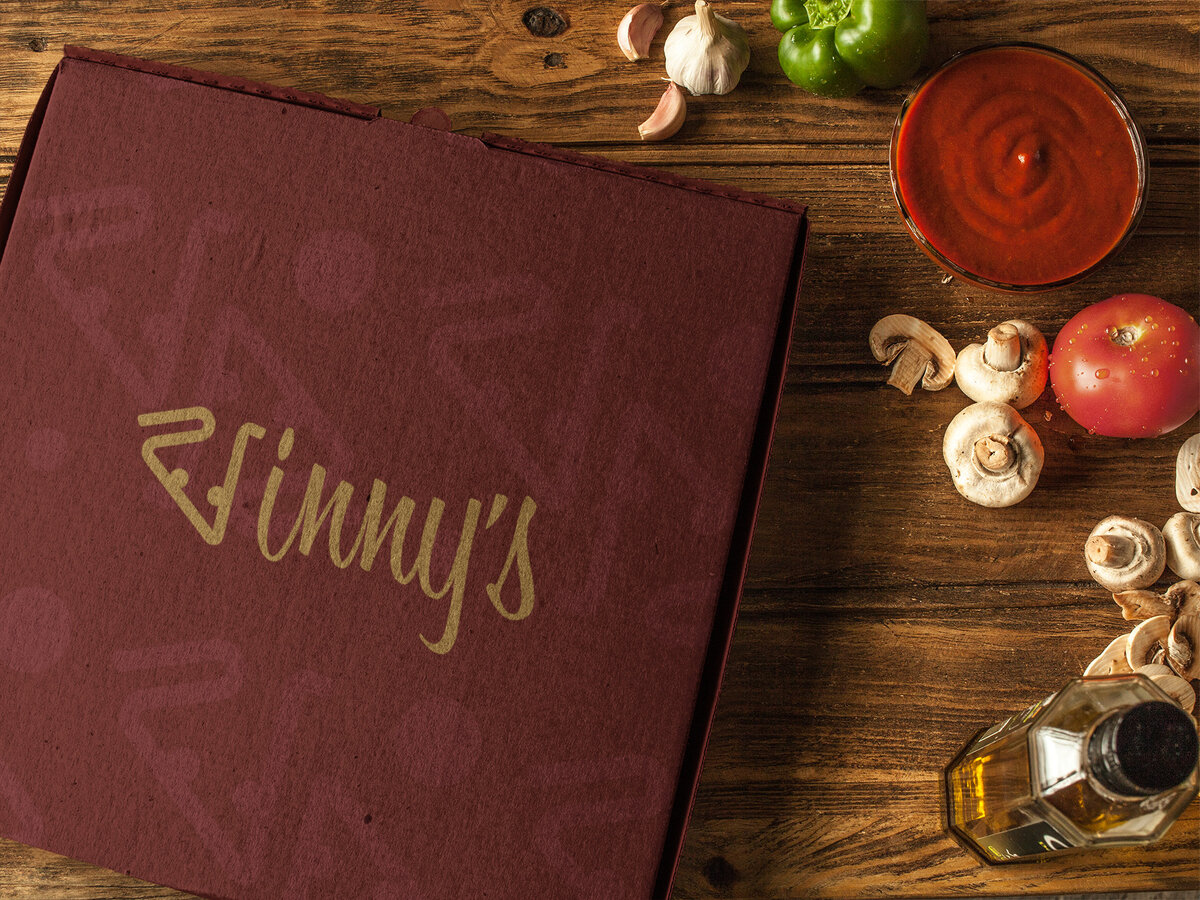 A Vinnys pizza box designed by Ayanna Richilano Designs sitting on a wood table with pizza ingredients set around the upper right corner