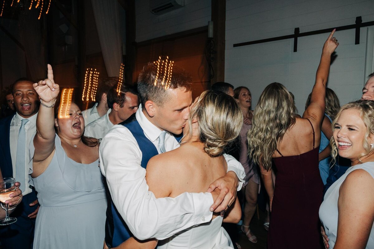 Couple kissing on the dance floor surrounded by joyful guests at a wedding reception.