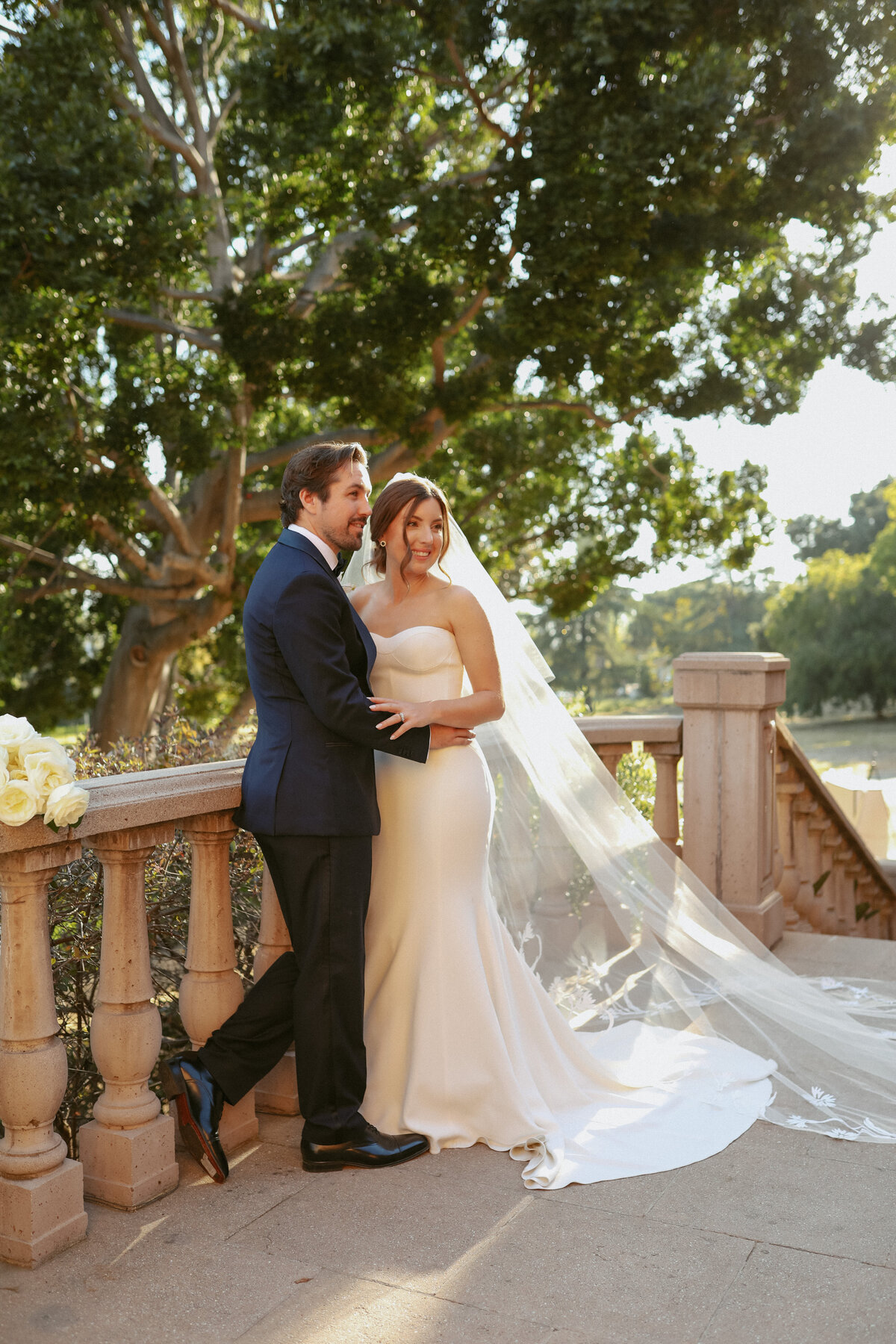 bride and groom standing against stone railing smiling at the camera with big green tree in the back round