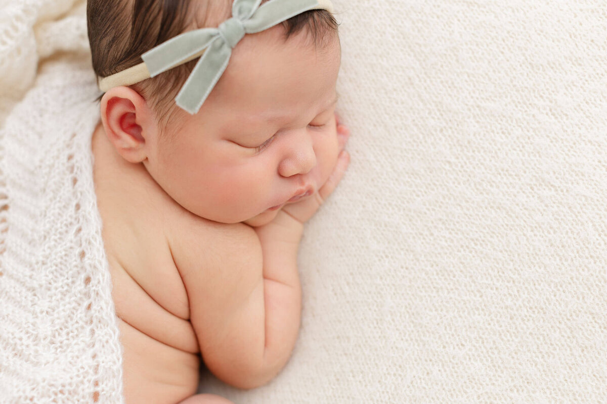baby sleeping on belly with hand under face. Baby is wearing a green velvet bow with a textured beige knit blanket draped across her back.