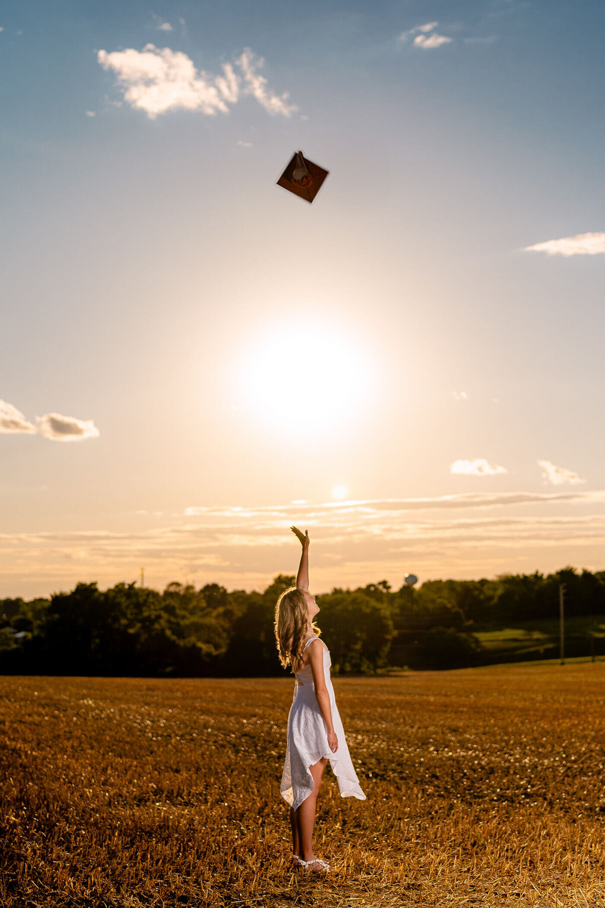 Senior portrait of a college grad tossing her cap into the air in a field at sunset