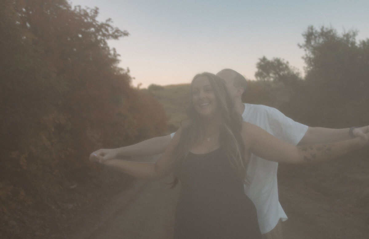 A man holding his girlfriend's hands out to the sides. They are outdoors with the Fall colors around them.